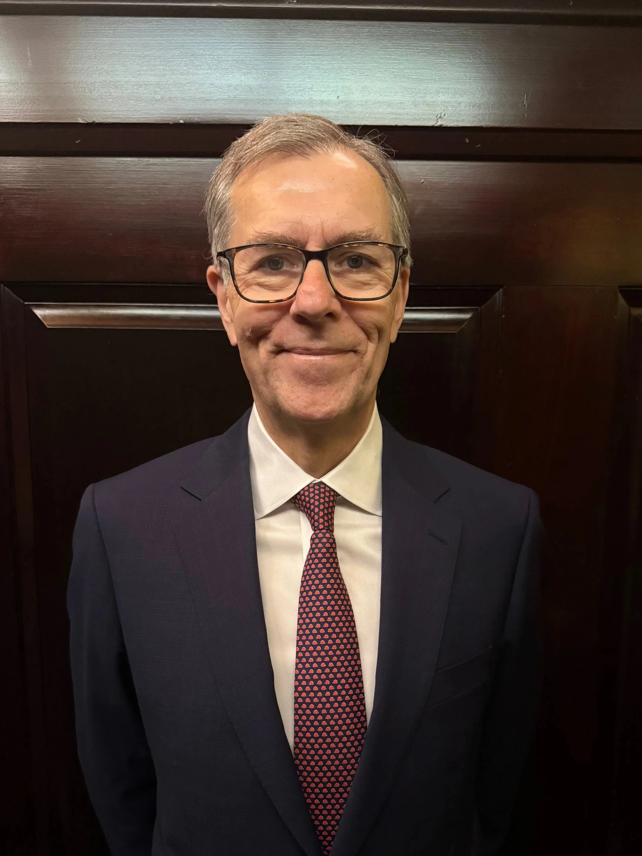 An Advisor dressed in a dark suit, white shirt, and red patterned tie, standing in front of a dark wooden background.