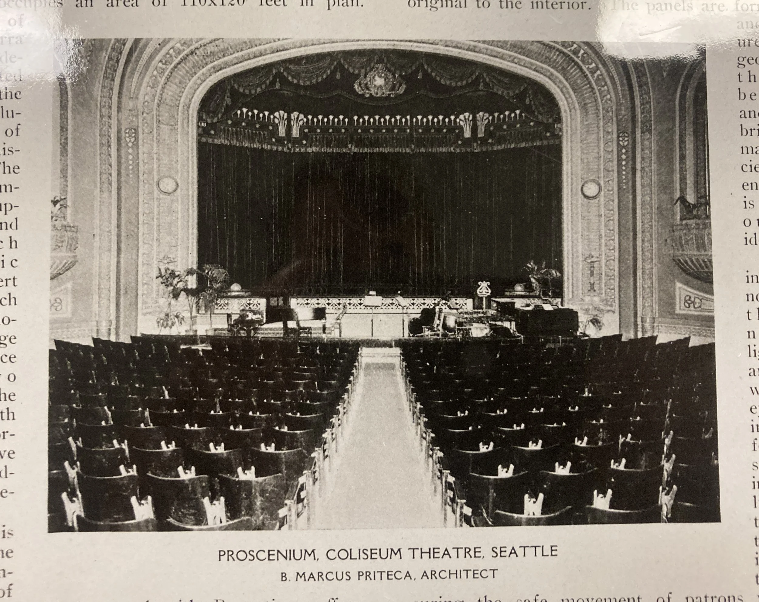 Black and white photograph of an empty theater stage with closed curtains, ornate arches, and seating area in the foreground.