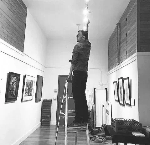 A person standing on a ladder inside an art gallery, observing framed artwork on the wall.