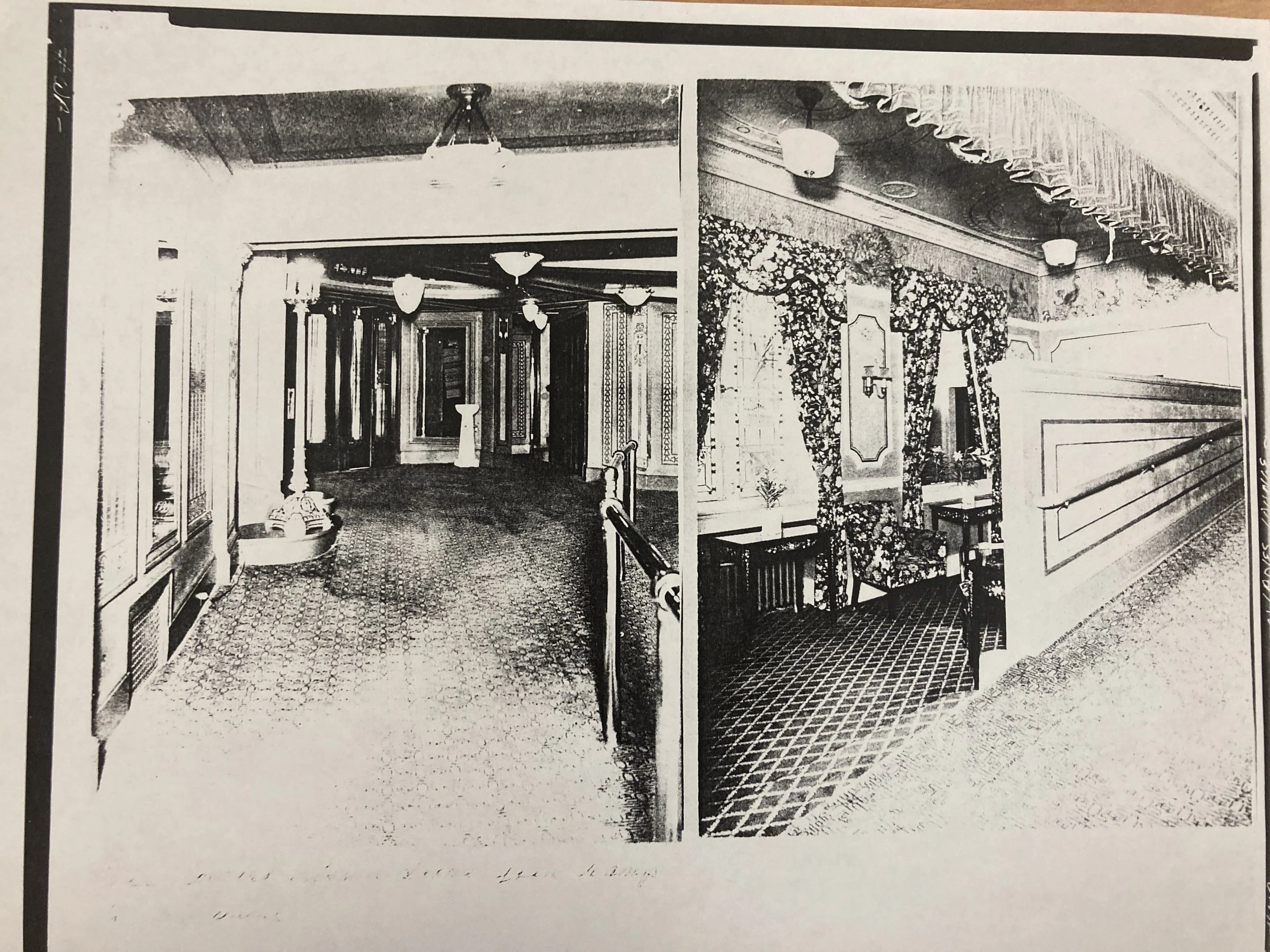 Black and white photograph of an ornate interior hallway with decorative lighting fixtures, patterned flooring, and detailed woodwork and wallpaper on the walls.