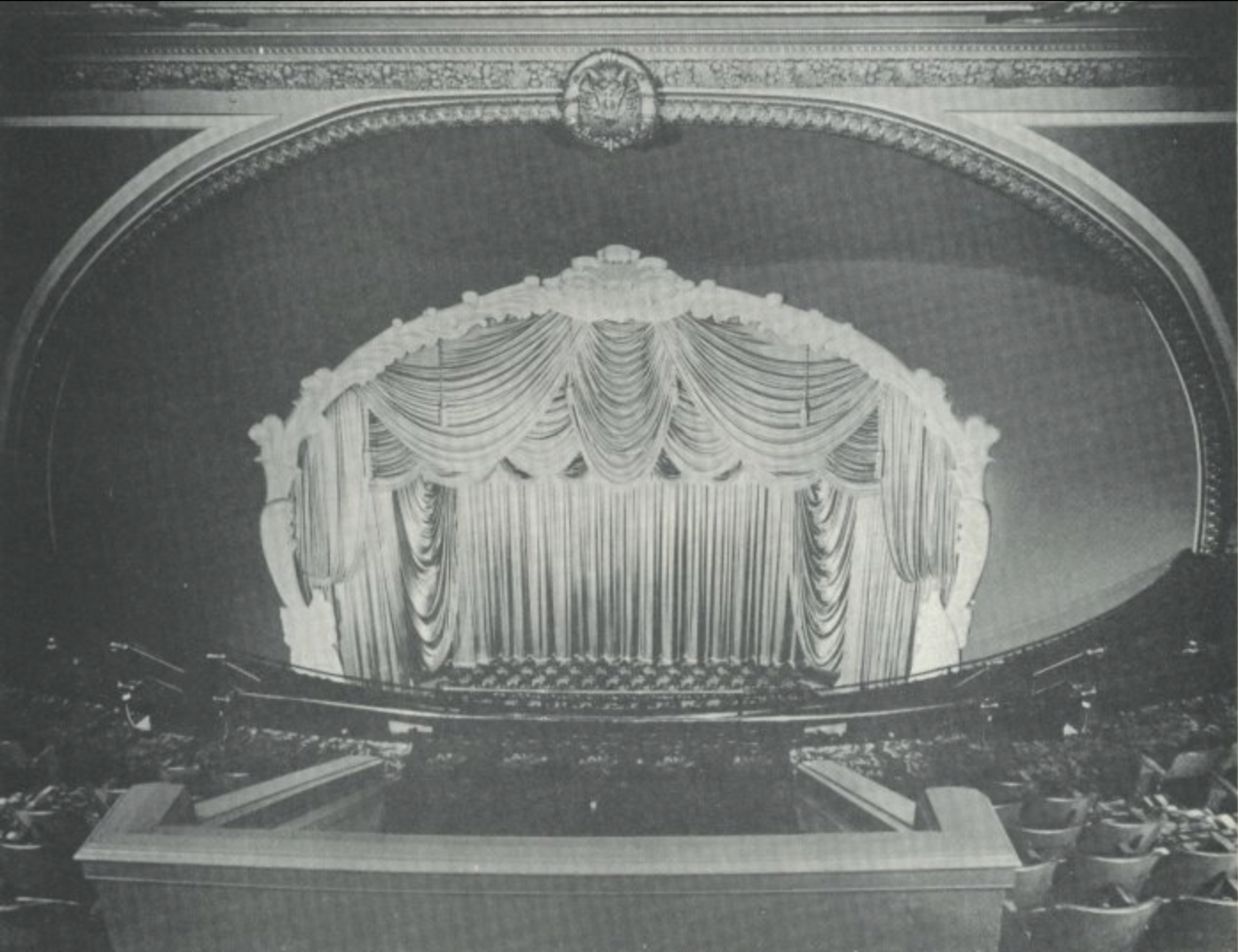 An empty theater stage with decorative curtains and ornate arch, viewed from the auditorium. The stage is set for a performance.