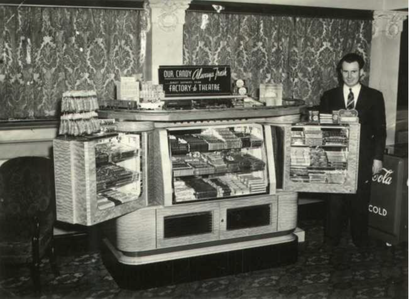 Black and white photo of a vintage candy and snack display case with an employee standing beside a Coca-Cola refrigerated unit, featuring a sign that reads 'Our Candy Always Fresh' in a retro setting.