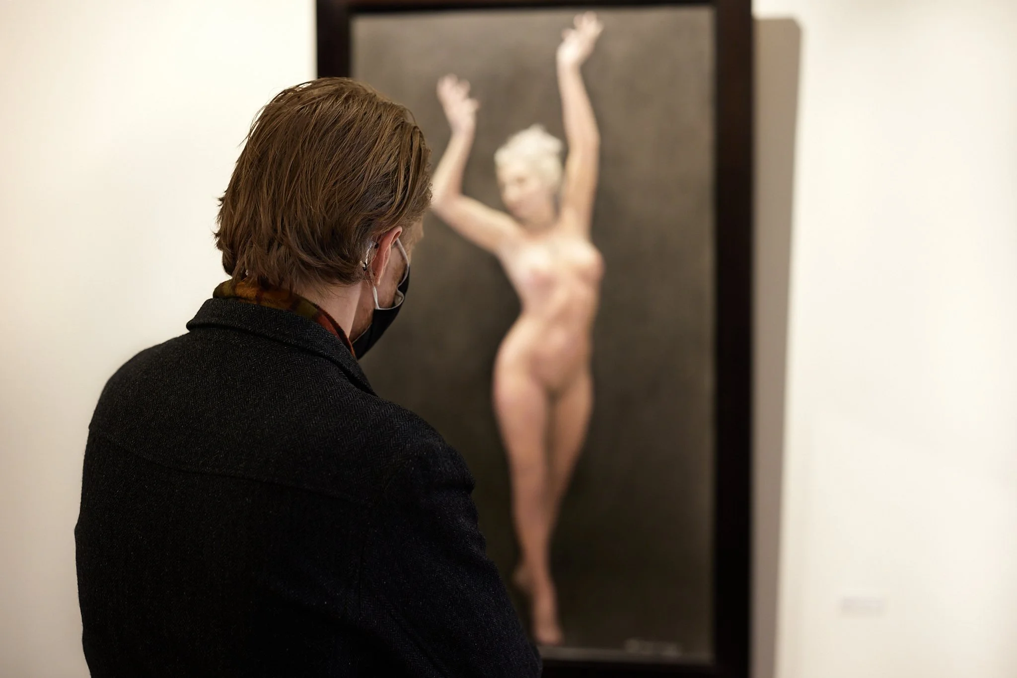 Person with brown hair and black face mask observing a nude female mannequin with arms raised, in an art gallery.