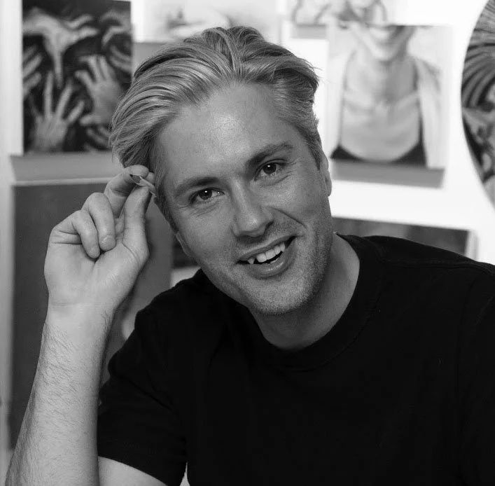 Black and white photograph of a man smiling, with hairstyled hair, wearing a black t-shirt, sitting indoors with artwork in the background.