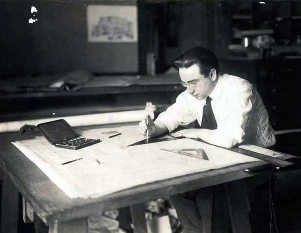 A man in a white shirt and black tie working on a large drafting table with drawing tools and a calculator nearby.