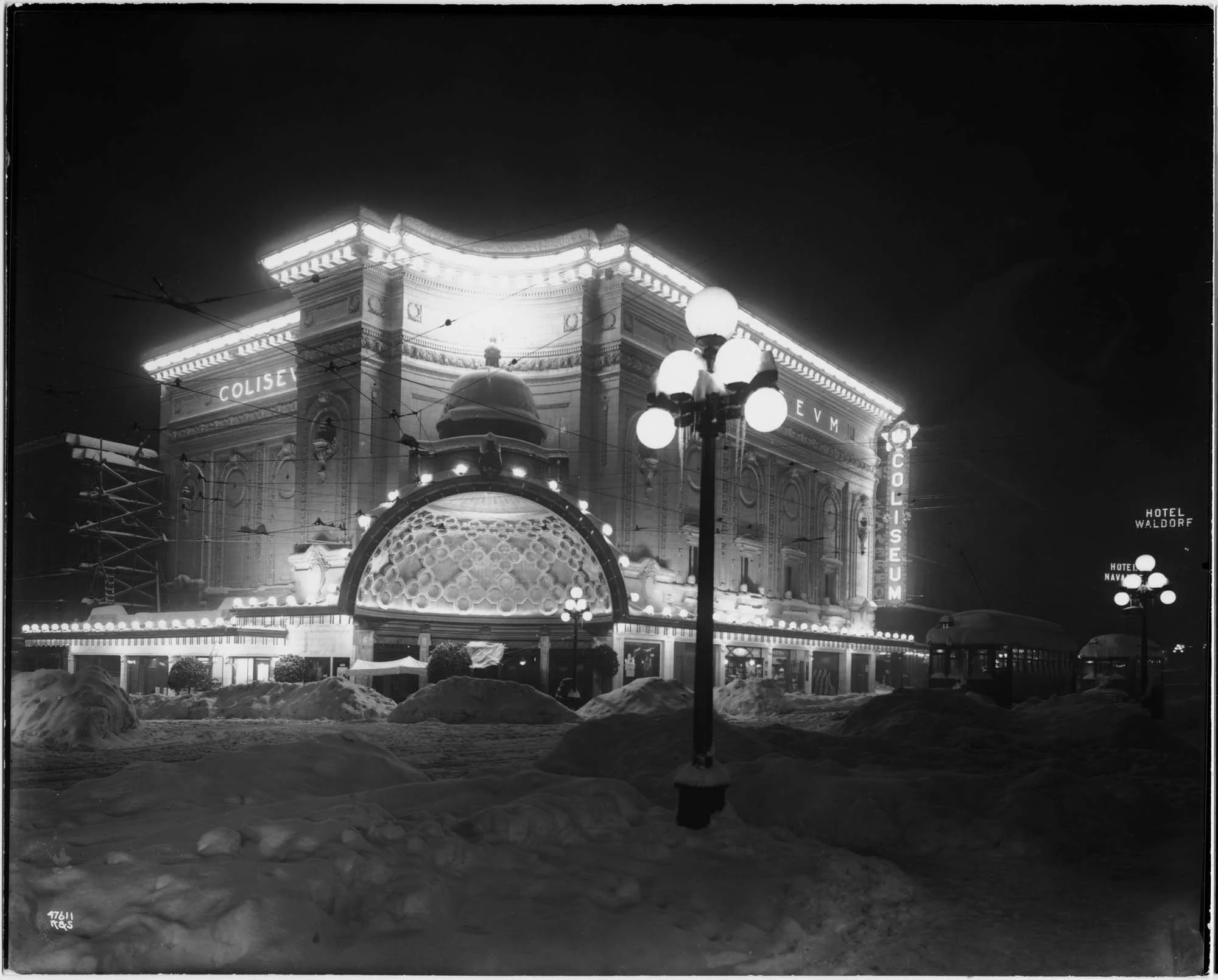 Nighttime scene of a historic building with neon lights reading 'Coliseum,' illuminated architectural details, and a sign for 'Hotel Waldorf' visible on the right side. Snow-covered ground and a street lamp in the foreground.