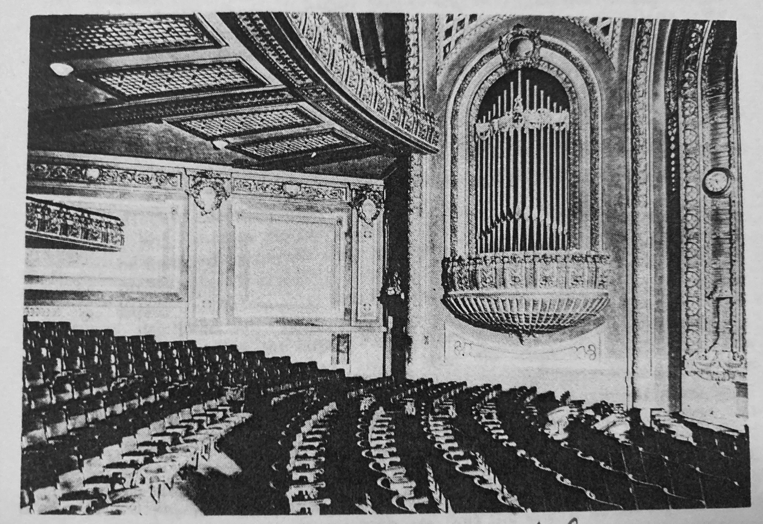 A historical photograph of a theater with ornate woodwork, high vaulted ceiling, and a large pipe organ in the background.