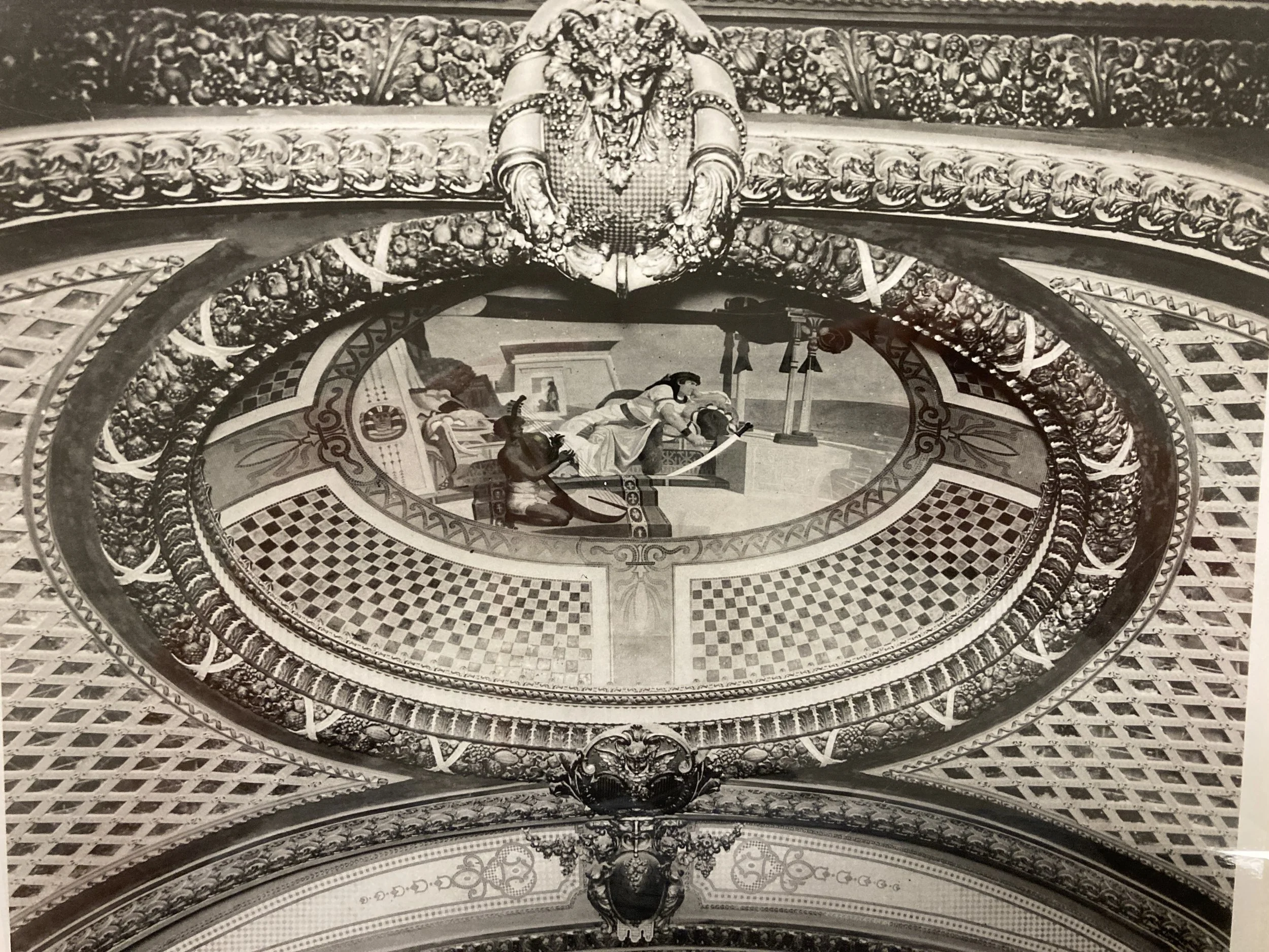 A black and white photo of an ornate ceiling with detailed artwork, including a scene with figures, decorative borders, and a central face motif at the top.