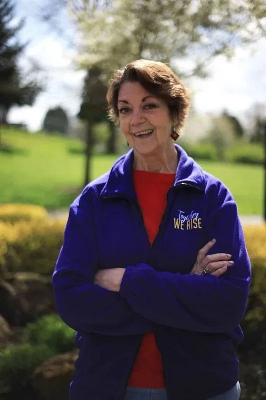 A woman with short brown hair smiling outdoors, wearing a purple jacket with 'Together We Rise' embroidered on it, over a red shirt, standing with arms crossed in a park with trees and grass in the background.