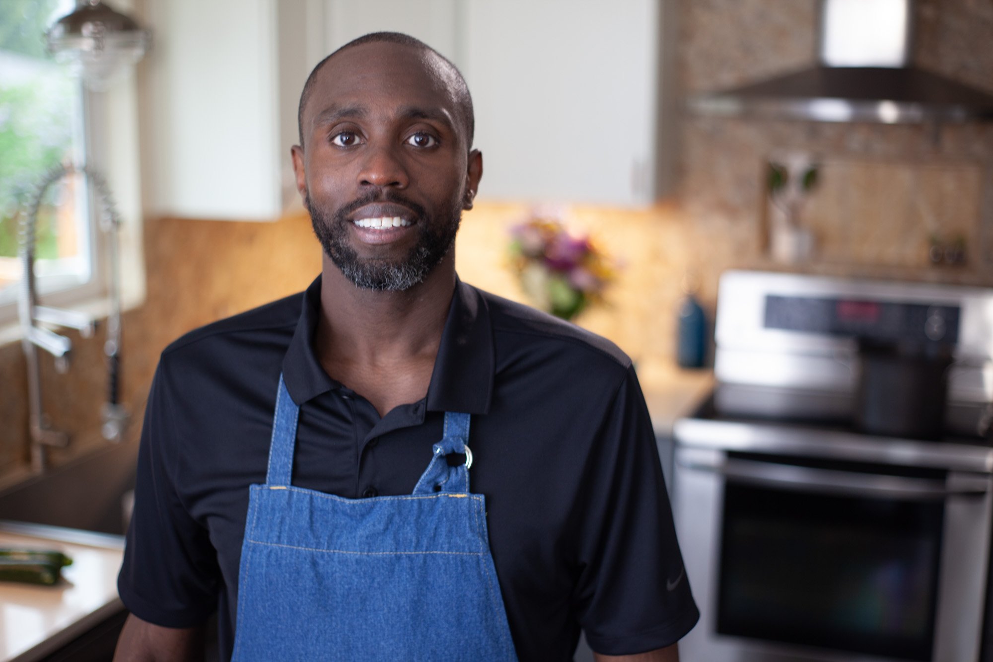 Smiling man with a beard and short hair wearing a black shirt and a blue apron in a kitchen.
