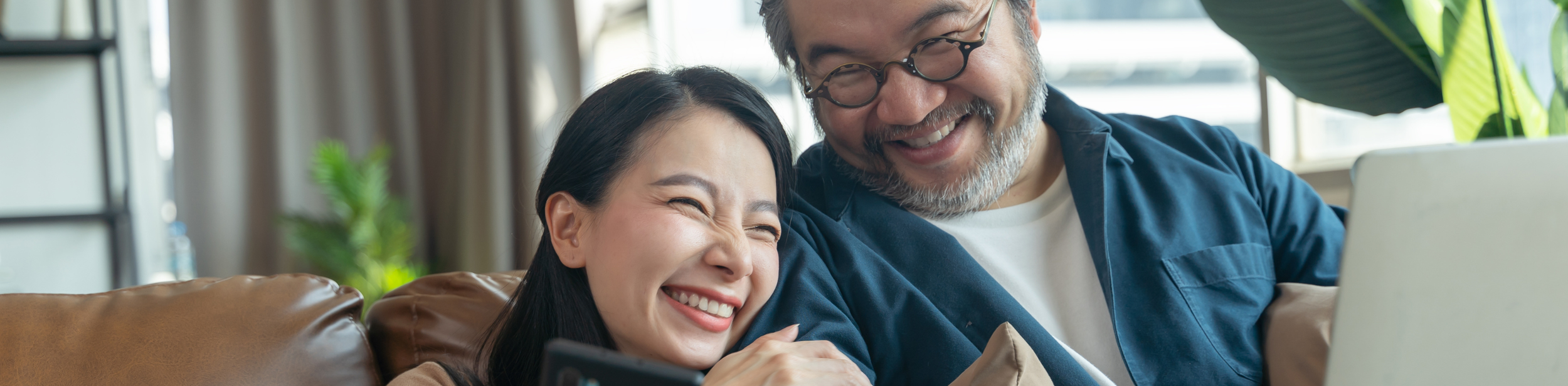 A smiling woman and man sitting on a couch, looking at a laptop and phone together in a bright room.