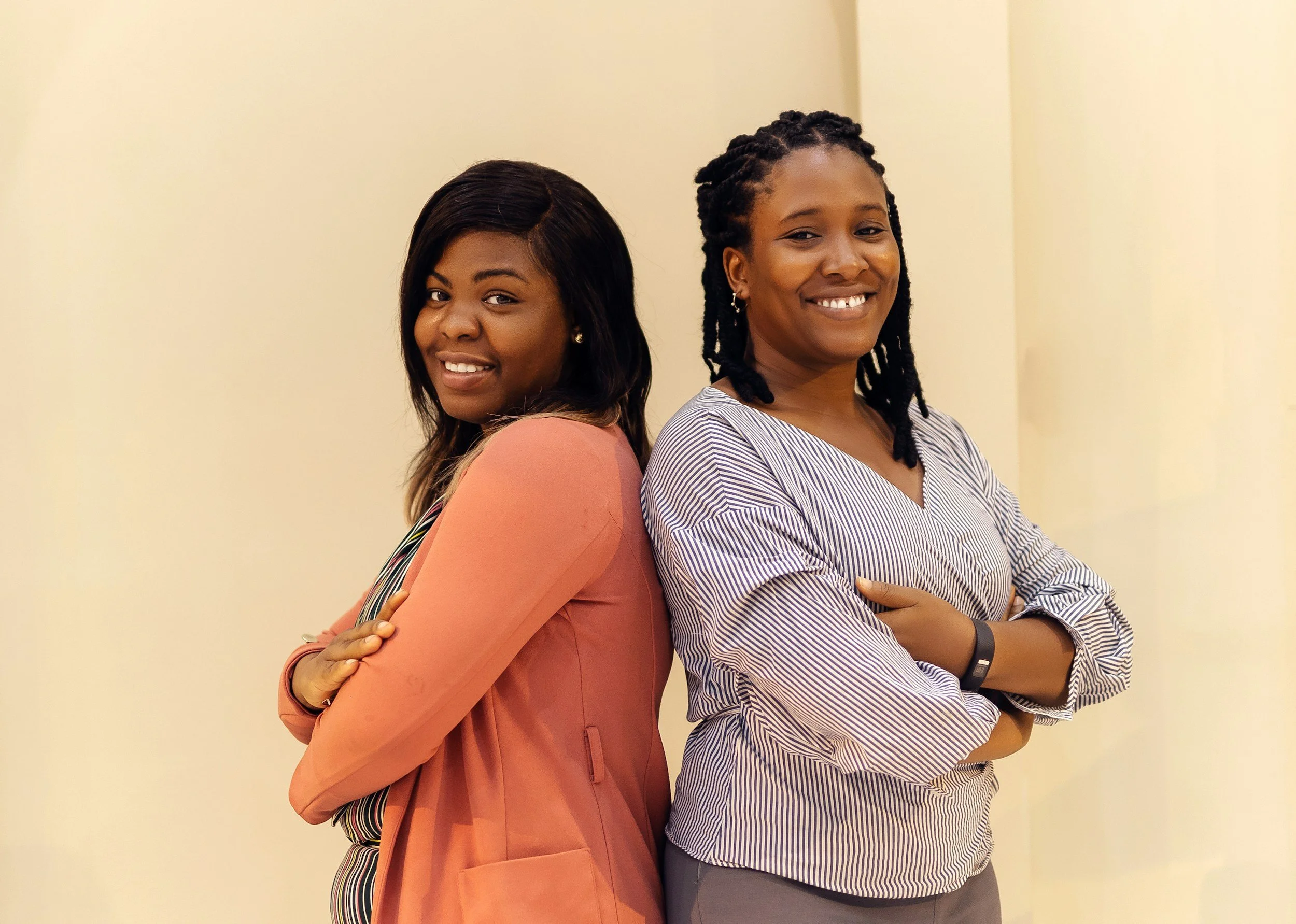 Two women standing back-to-back, smiling at the camera, with arms crossed in front of a plain light cream-colored wall.