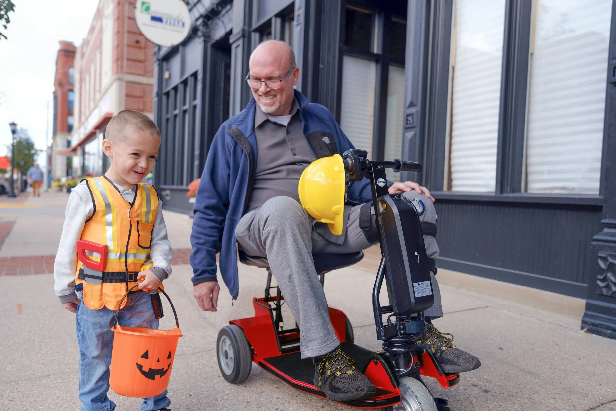 A man with a bald head, glasses, and a gray shirt smiles while sitting on a mobility scooter. A young boy dressed in a Halloween pumpkin-themed vest holds a bright orange pumpkin-shaped bucket for trick-or-treating. They are on a city sidewalk, with buildings and pedestrians visible in the background.