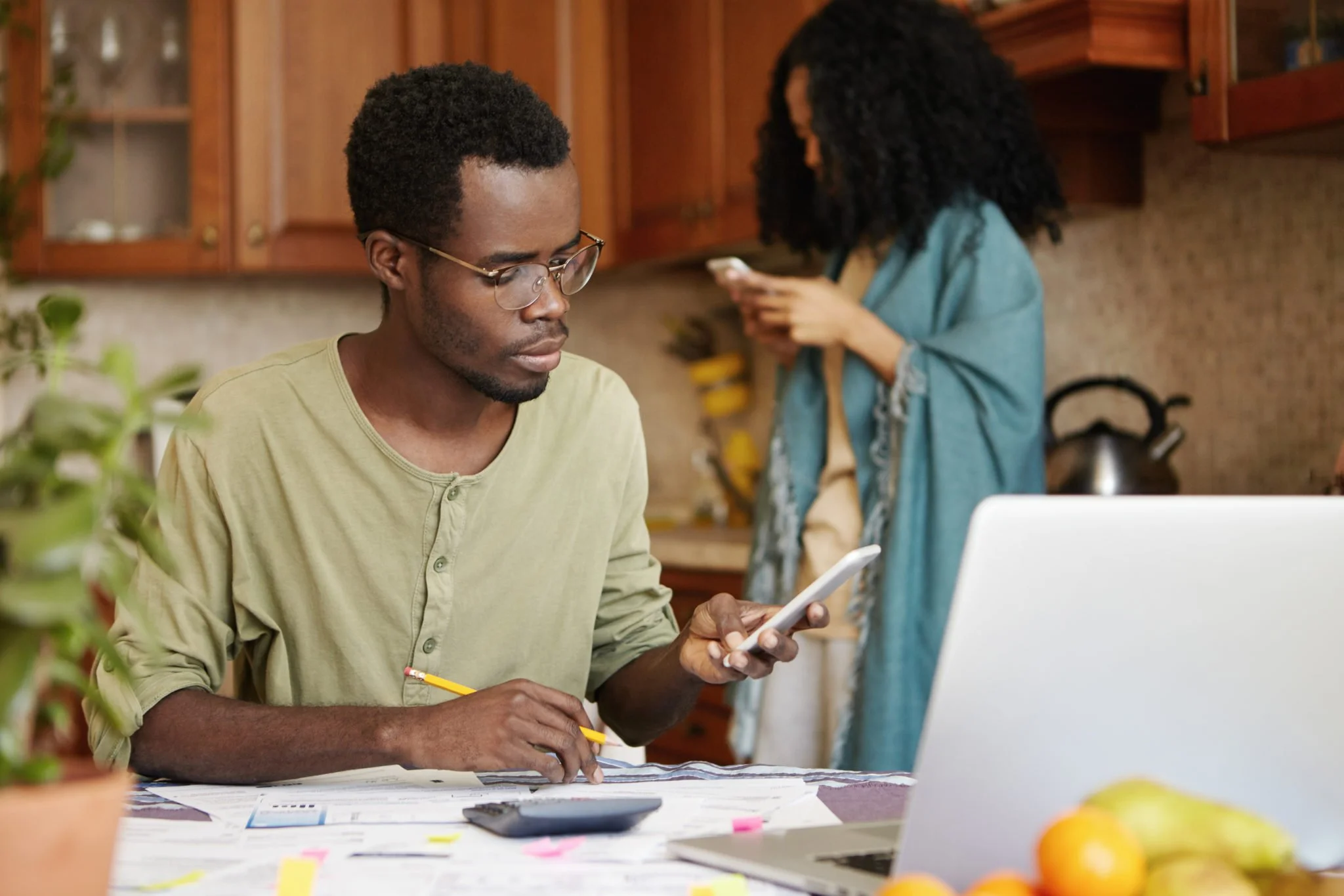 A young man with glasses sitting at a table using a smartphone, with a woman in the background in a kitchen looking at her phone.