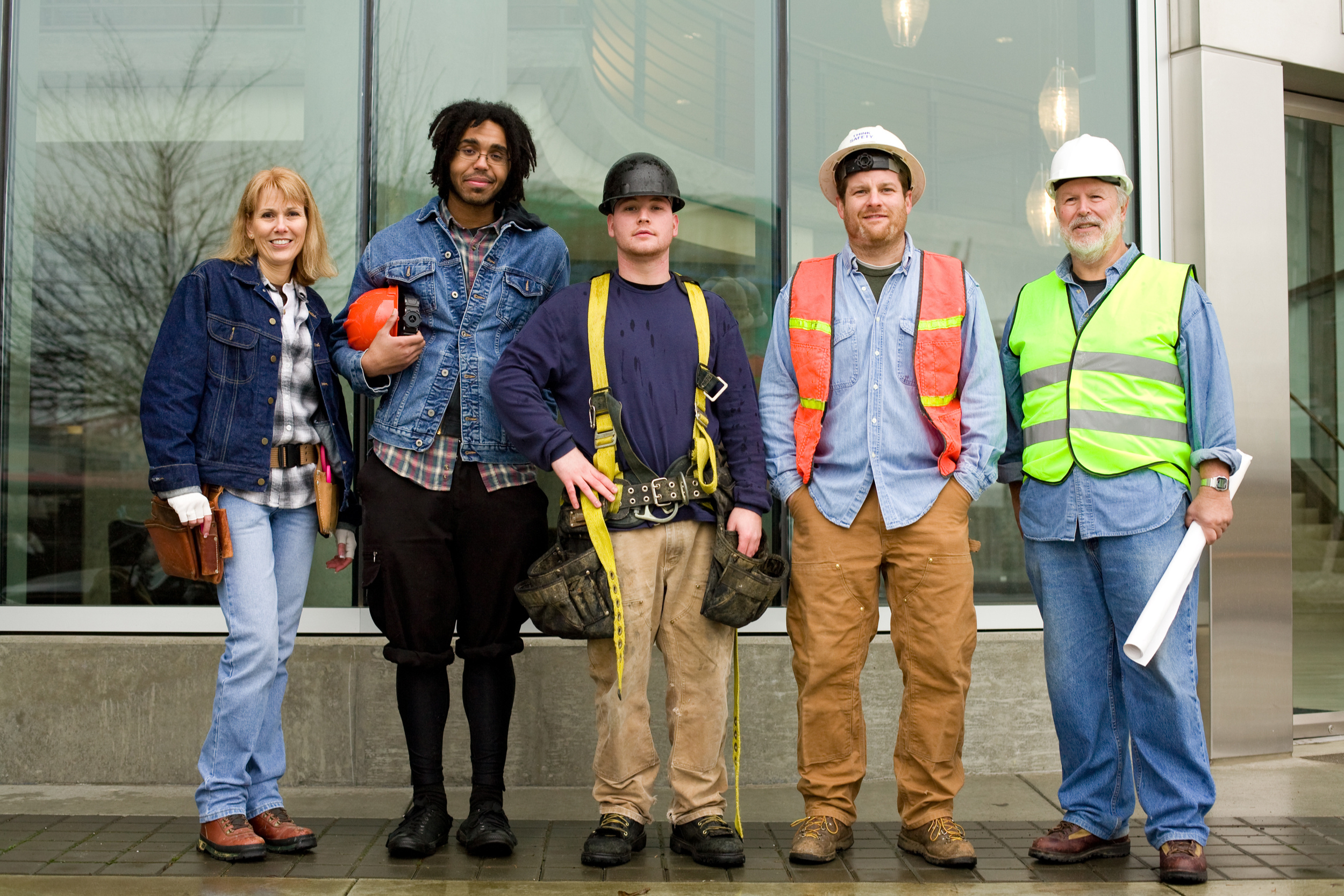 Group of five construction workers standing outside a glass building, smiling and looking at the camera. They are wearing safety gear including helmets, vests, and work boots.