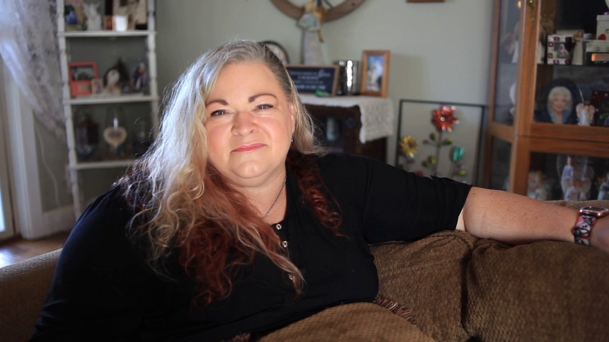 A woman with long, curly, blonde hair with reddish tips, sitting on a brown couch in a living room, smiling subtly at the camera.