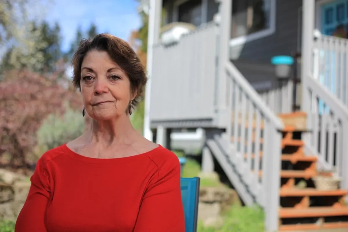 Older woman with short brown hair and wearing a red top standing outdoors in a backyard with a house and stairs in the background.