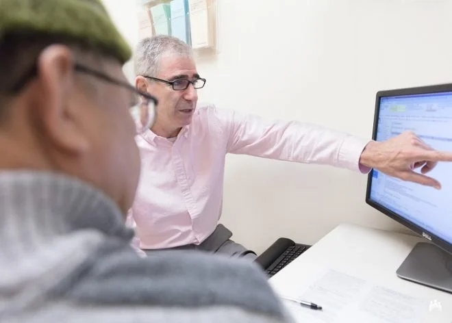Two men in an office, one wearing glasses and a pink shirt and the other with glasses and a gray hoodie, looking at a computer screen where the man in the pink shirt is pointing at information displayed on the monitor.