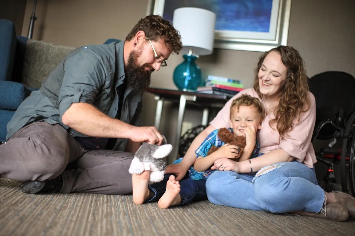 Family sitting on the floor playing with a stuffed animal, a man, woman, and young boy smiling, cozy living room setting.