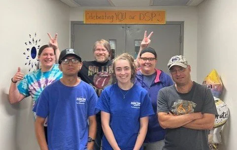 Group of five people standing in front of a door with a sign that says "Celebrating YOU a DSP". Some are smiling; one person is giving a thumbs-up, and another is making a peace sign. The group appears to be celebrating or acknowledging an event or achievement.