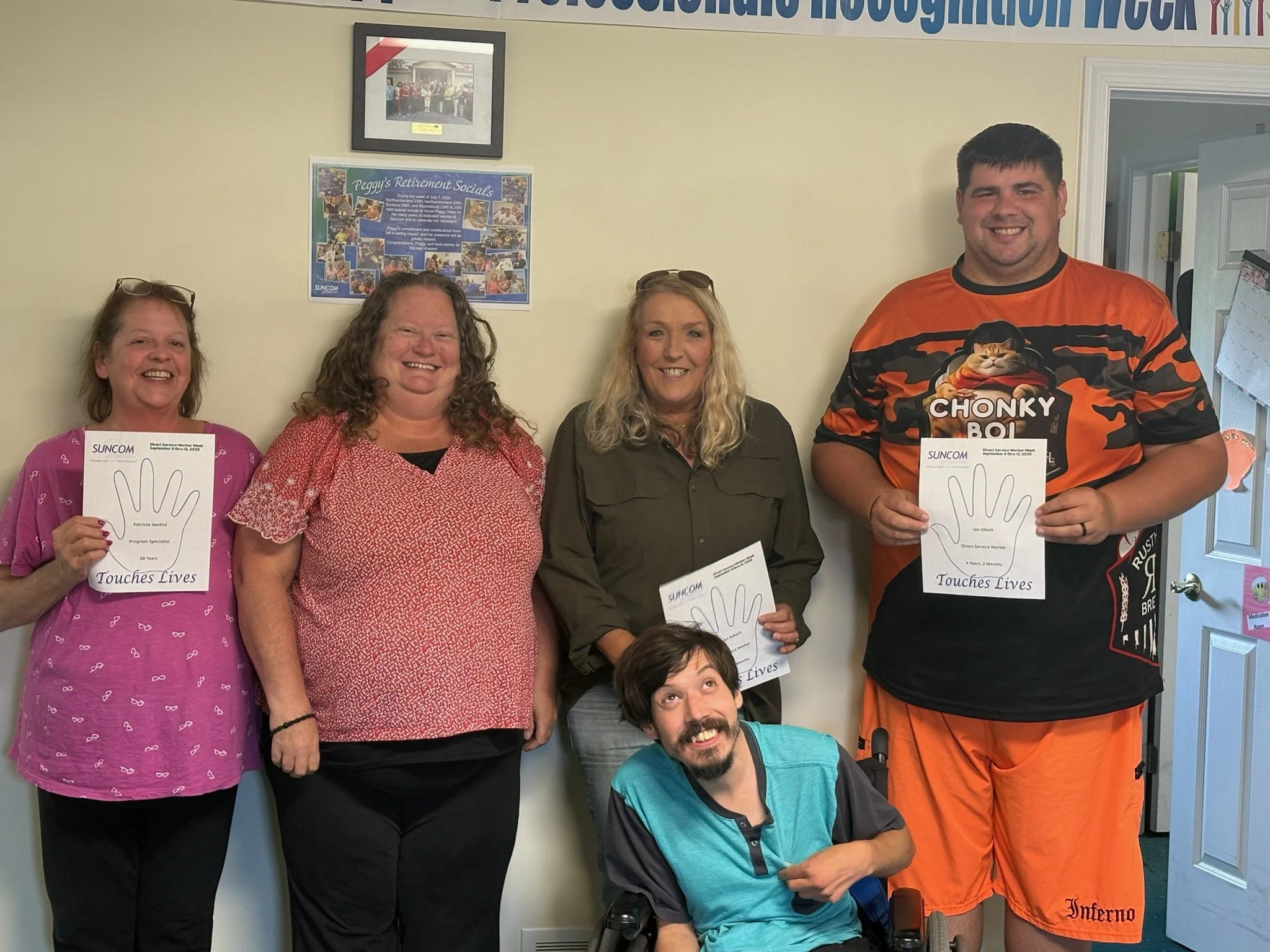 Group of five people, four women and one man, standing indoors and holding signs for a program. The woman on the far left wears glasses and a pink shirt, the second woman wears a red patterned shirt, the third woman has blonde hair and wears a dark shirt, and the man is tall, wearing a black and orange sports shirt with a cat graphic. The fifth person is a man sitting in a wheelchair in front, smiling and pointing at his sign.