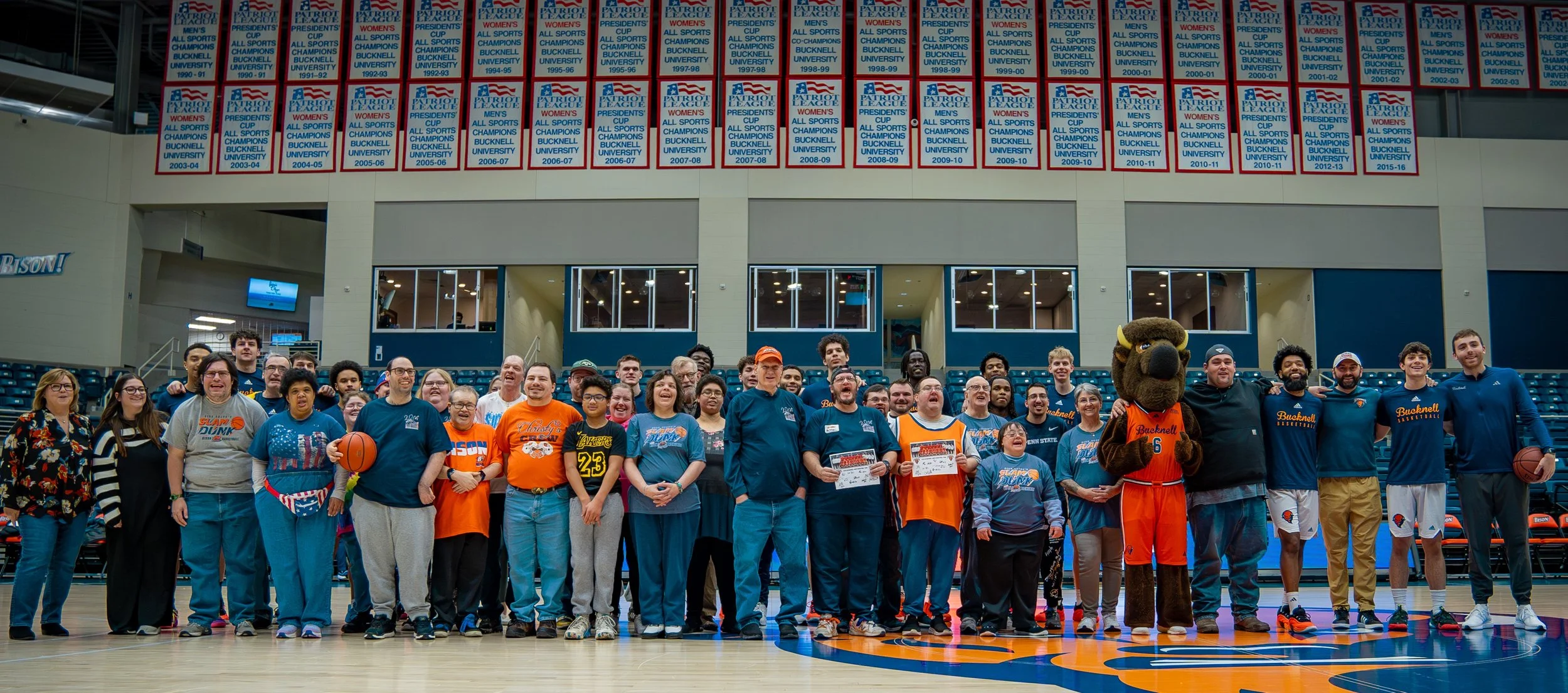 20th Annual Dirk Sojka's Slam Dunk with Bison Basketball - Jan 2025 A group photo of students, staff, and a mascot on a basketball court at Bucknell University, with banners hanging on the wall noting athletic championships.