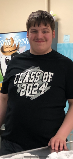 Young man with short dark hair and mustache wearing a black t-shirt with 'Class of 2024' printed on it, standing indoors.