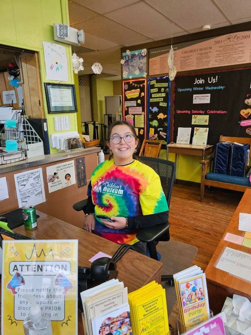 A young girl wearing a tie-dye children's museum T-shirt sitting at a desk, smiling, with a green soda can nearby, inside a colorful community center or museum.