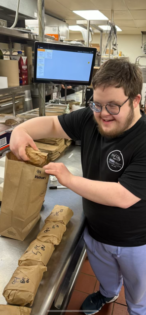 A young man with glasses smiling and packing brown paper bags in a kitchen or cafeteria setting.