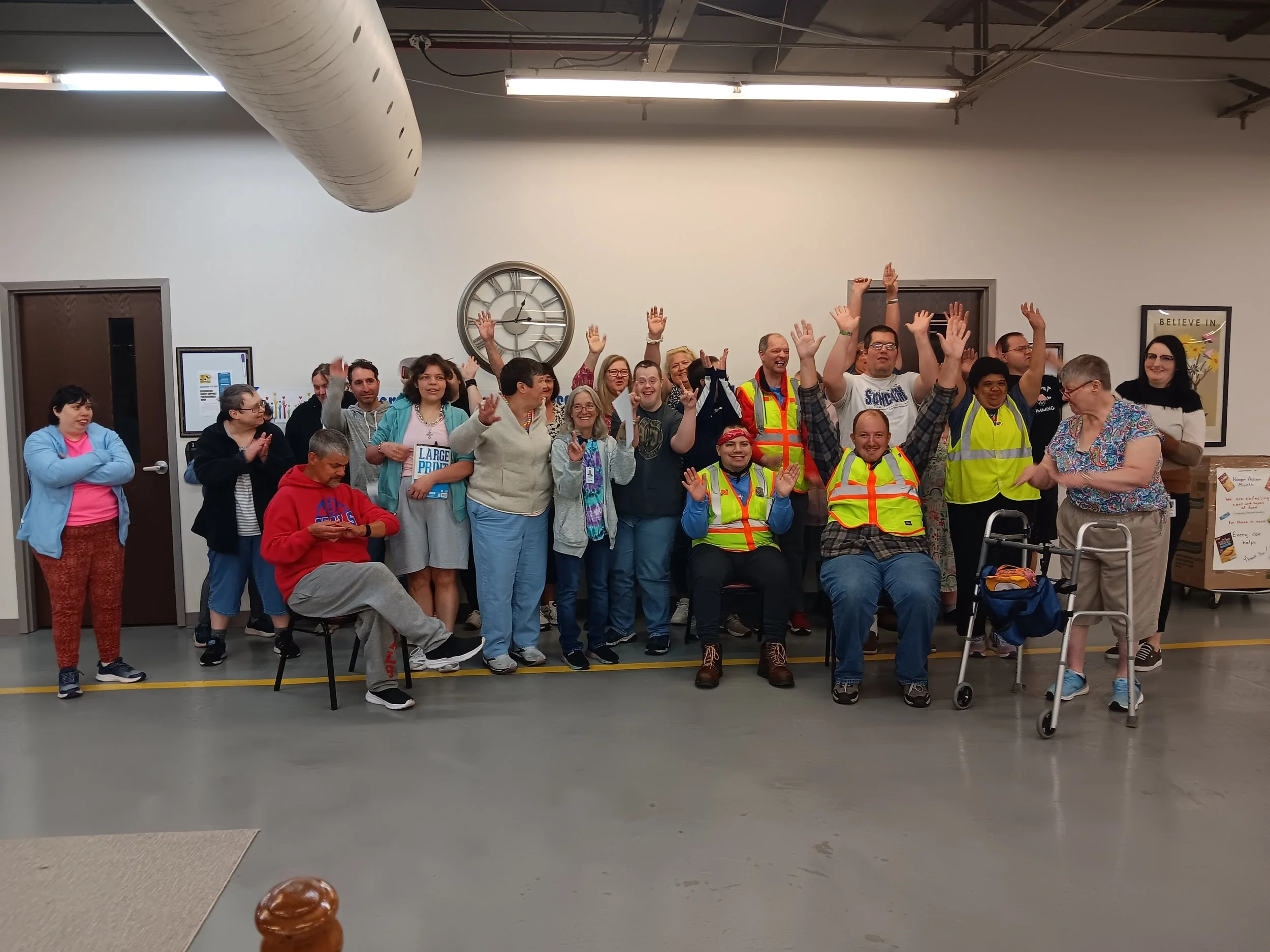 Group of diverse people gathered indoors, some wearing yellow safety vests, smiling and raising their hands in celebration. Northumberland Pre-vocational program.