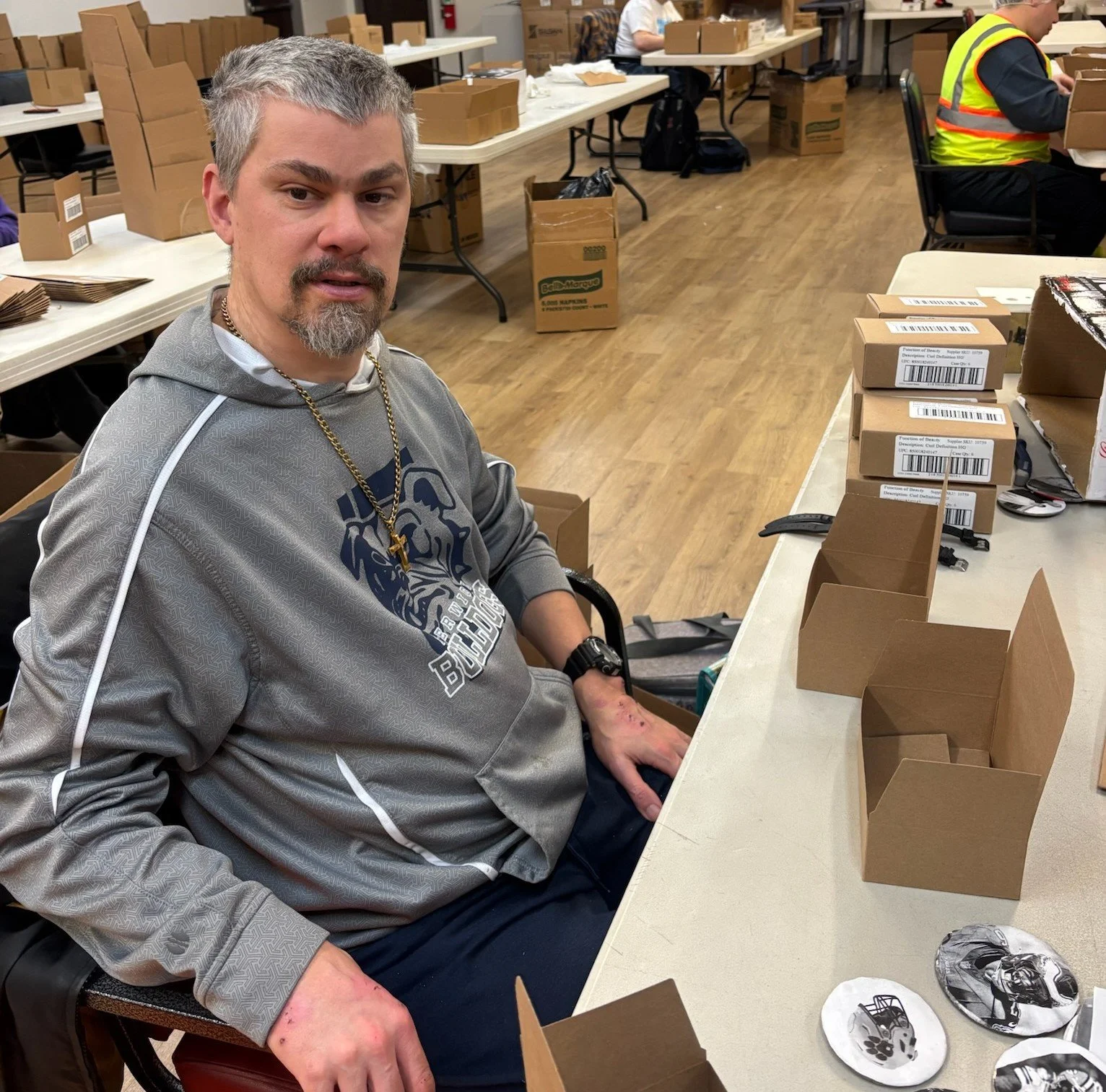 A man sitting at a table in a warehouse or packaging area, surrounded by cardboard boxes and packaging materials, with another person working in the background.