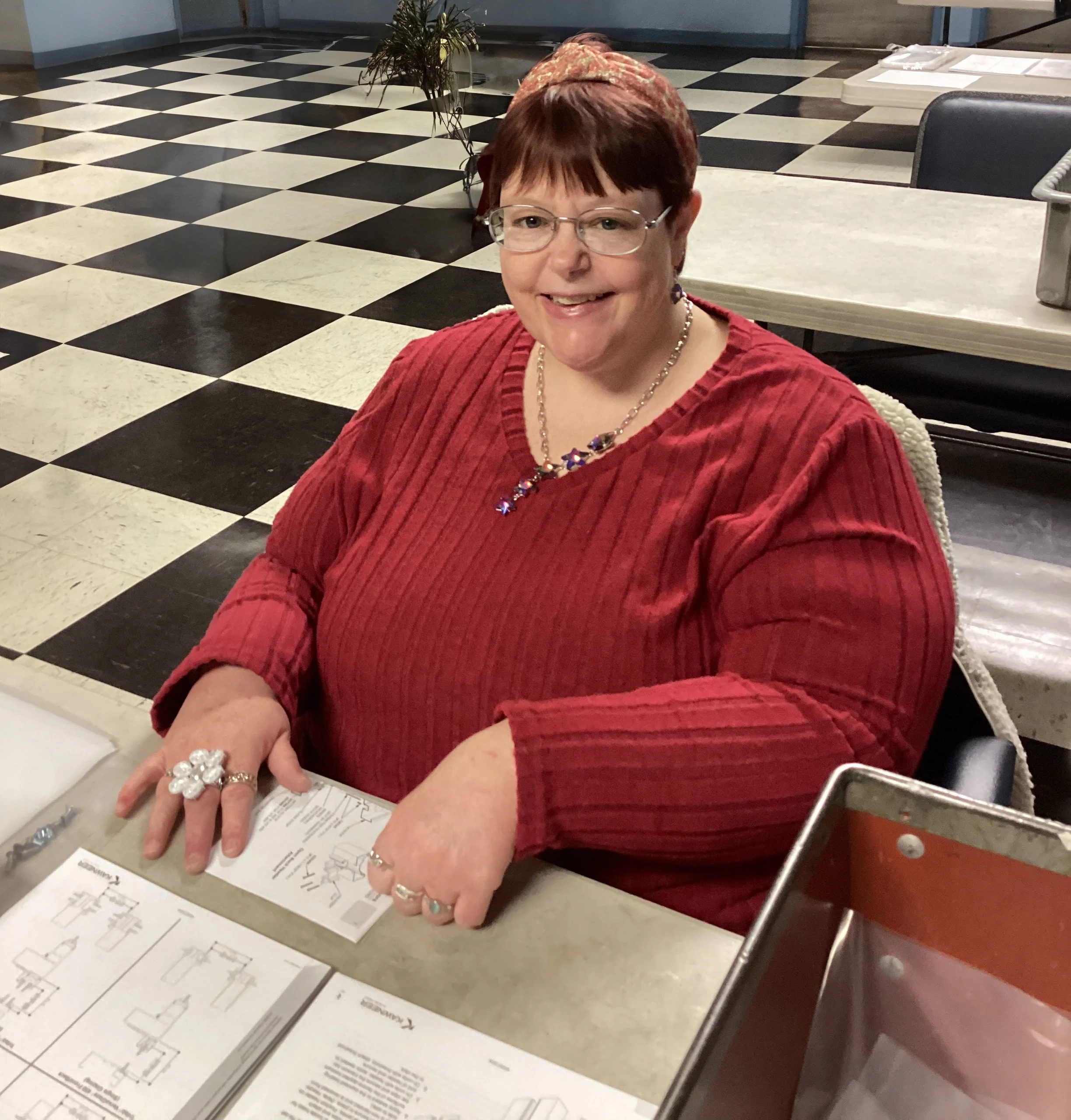 A smiling woman with short red hair, glasses, and pale skin, wearing a red long-sleeved top, sitting at a table with paperwork, in a room with black and white checkered floor and blue walls.