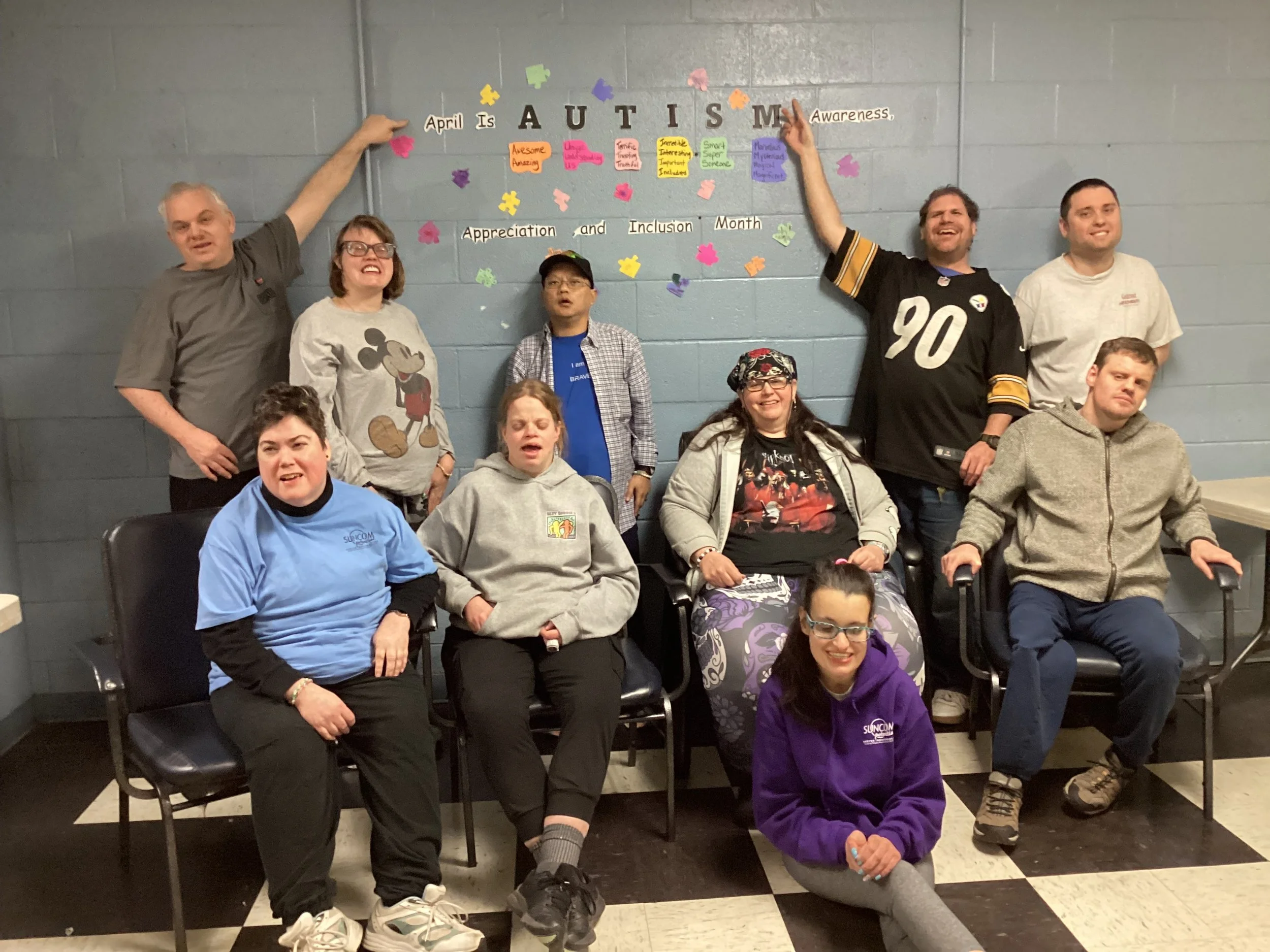 A group of ten diverse people, including men and women of different ages, poses together in front of a wall with colorful notes and decorations for Autism Awareness Month. Some are seated, others standing, with cheerful expressions.