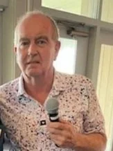 A man holding a microphone, wearing a patterned shirt, standing indoors near a window.