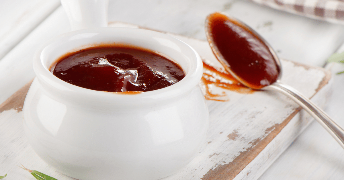 Close-up of a white bowl filled with dark syrup, with a silver spoon resting on its side and some syrup spilled on white wooden surface.