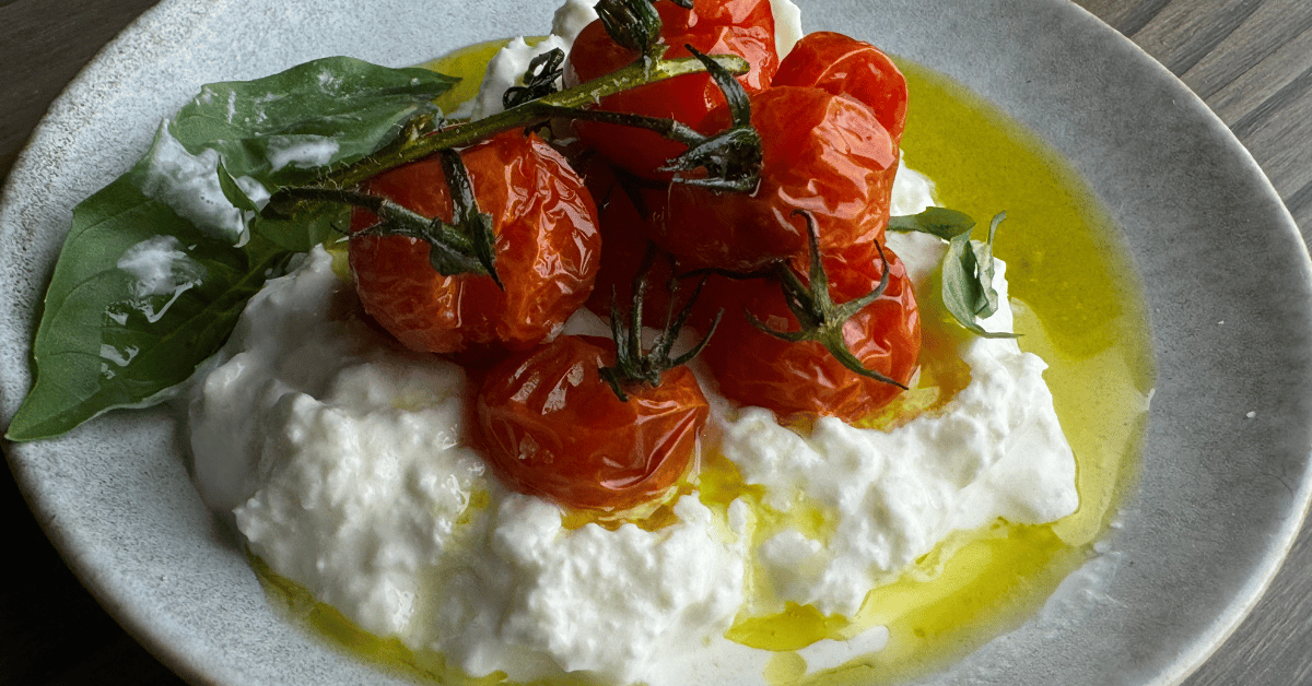 A plate of roasted cherry tomatoes on the vine, fresh basil leaves, ricotta cheese, and a drizzle of olive oil.
