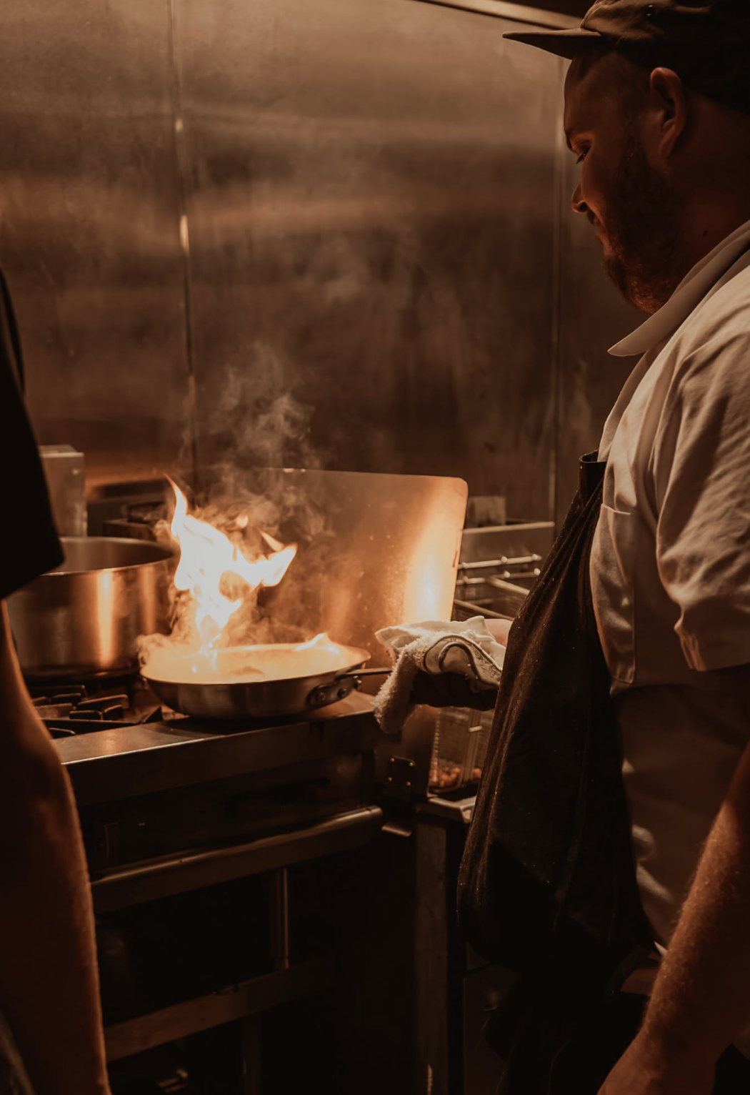 A chef in a white shirt and black apron is cooking on a stove with a fire flaring up in a professional kitchen.
