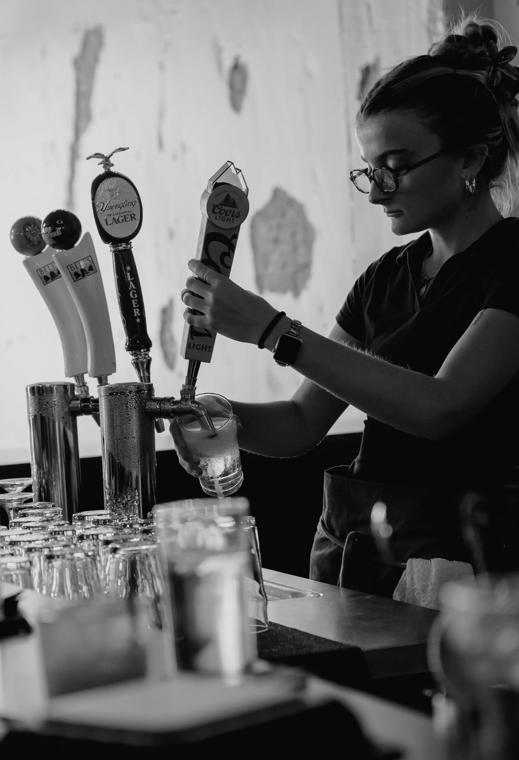 A woman with glasses and a hair bun wearing a dark shirt pours beer from multiple tap handles into a glass at a bar. The background features a distressed wall with peeling paint, and several glasses are on the bar in the foreground.