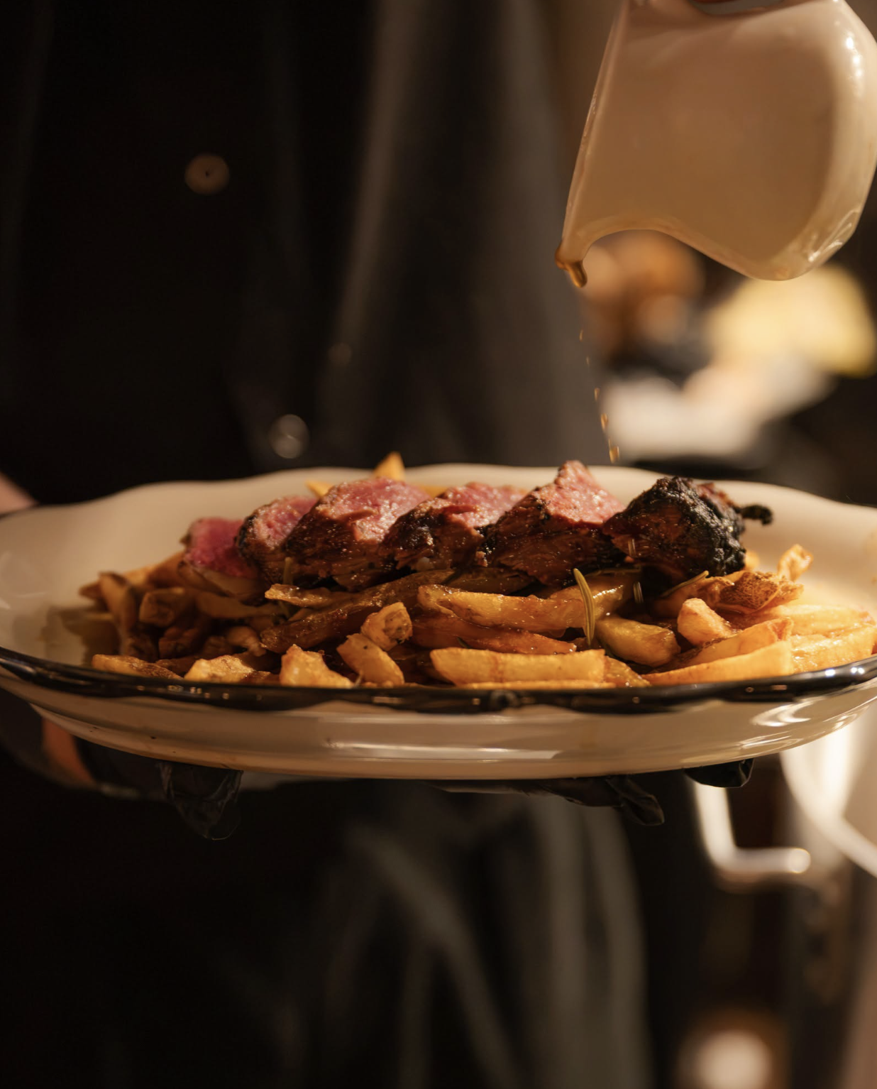 A chef is pouring creamy gravy over a plate of steak and fries.
