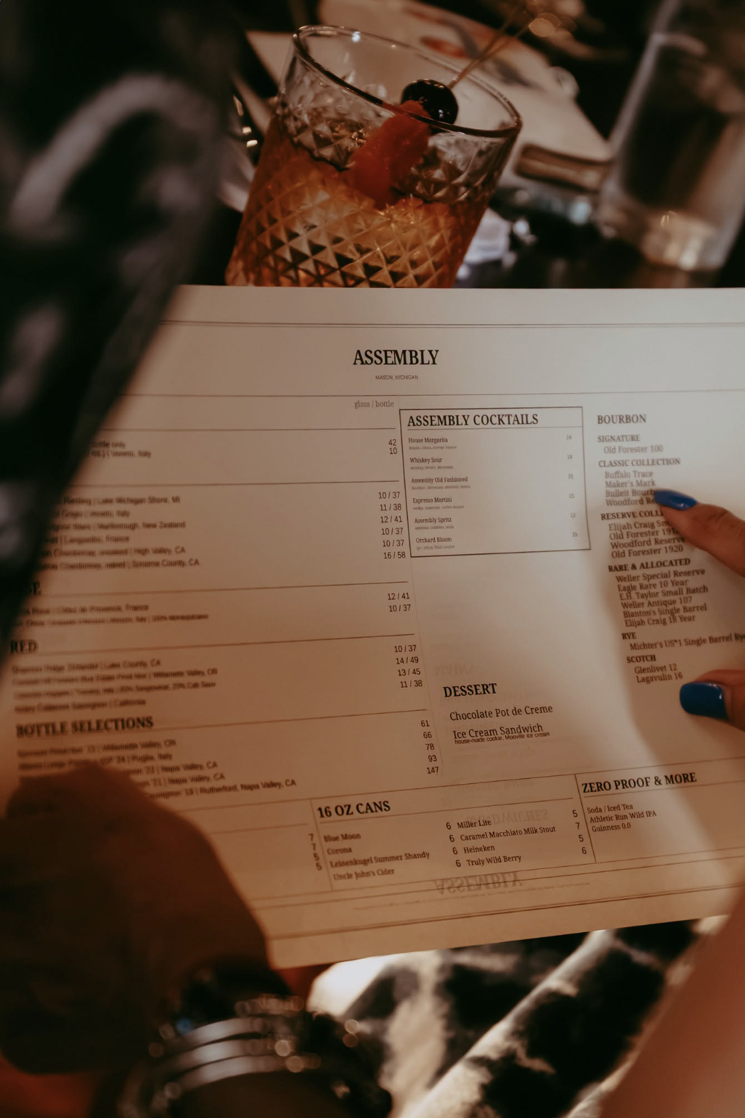 A person holding a menu with a cocktail drink and a drink glass on the table in the background.