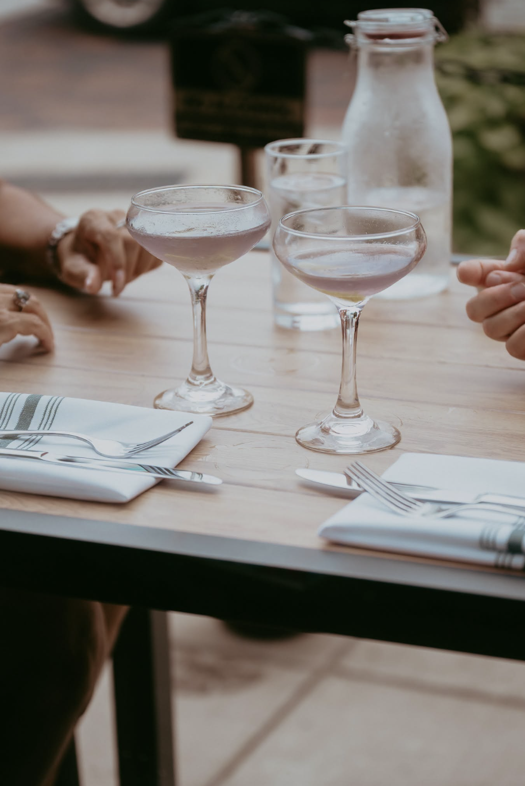 Two cocktails in coupe glasses on a wooden table with cloth napkins and silverware, with hands of people visible and a glass water bottle in the background.
