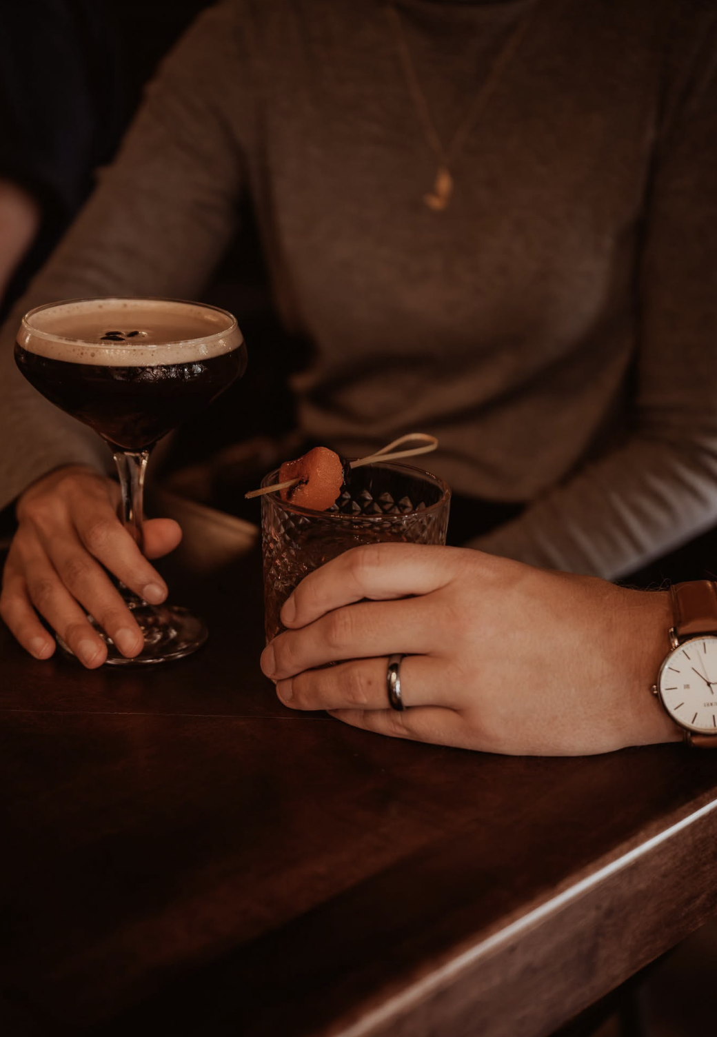 Two people holding cocktails at a bar. One person holds a dark-colored drink in a fancy glass, the other a reddish drink garnished with a piece of watermelon. Both are wearing rings, and one person has a watch.