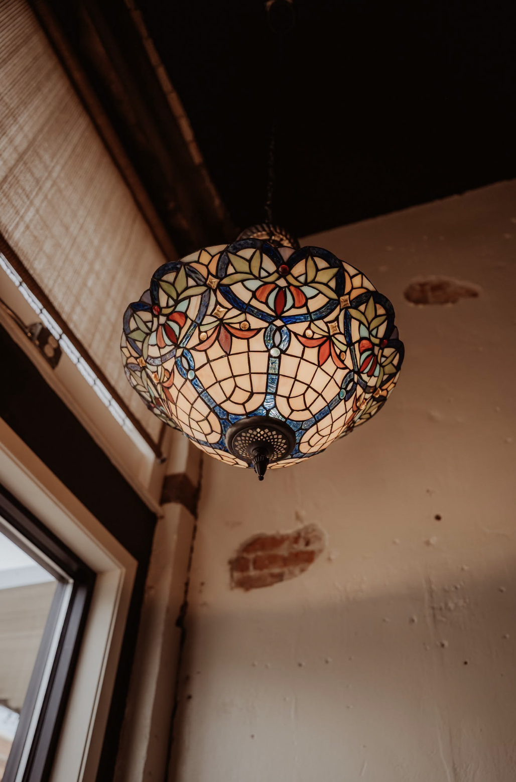 Colorful stained glass ceiling lamp hanging from ceiling in a room with brick wall and window.