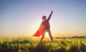 young girl in a field wearing a superhero cape while she thrusts an arm into the air