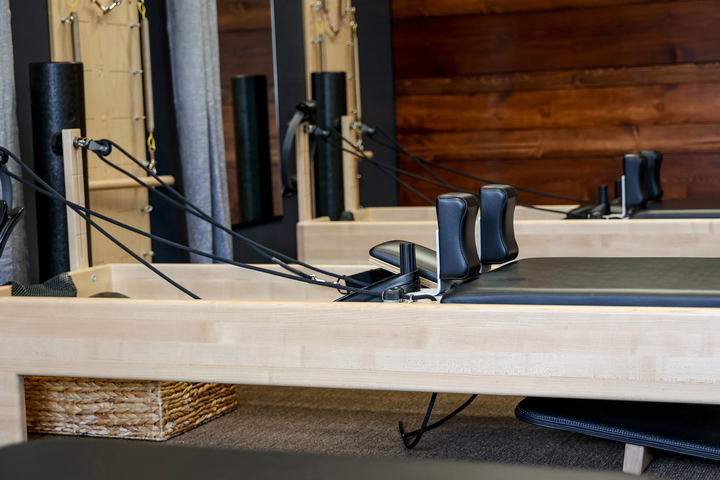 Interior of Collective Motion studio with black Pilates reformer equipment, wooden wall panels, a tall wooden wall rack with resistance bands, a wicker basket, a row of black chairs, and a table.