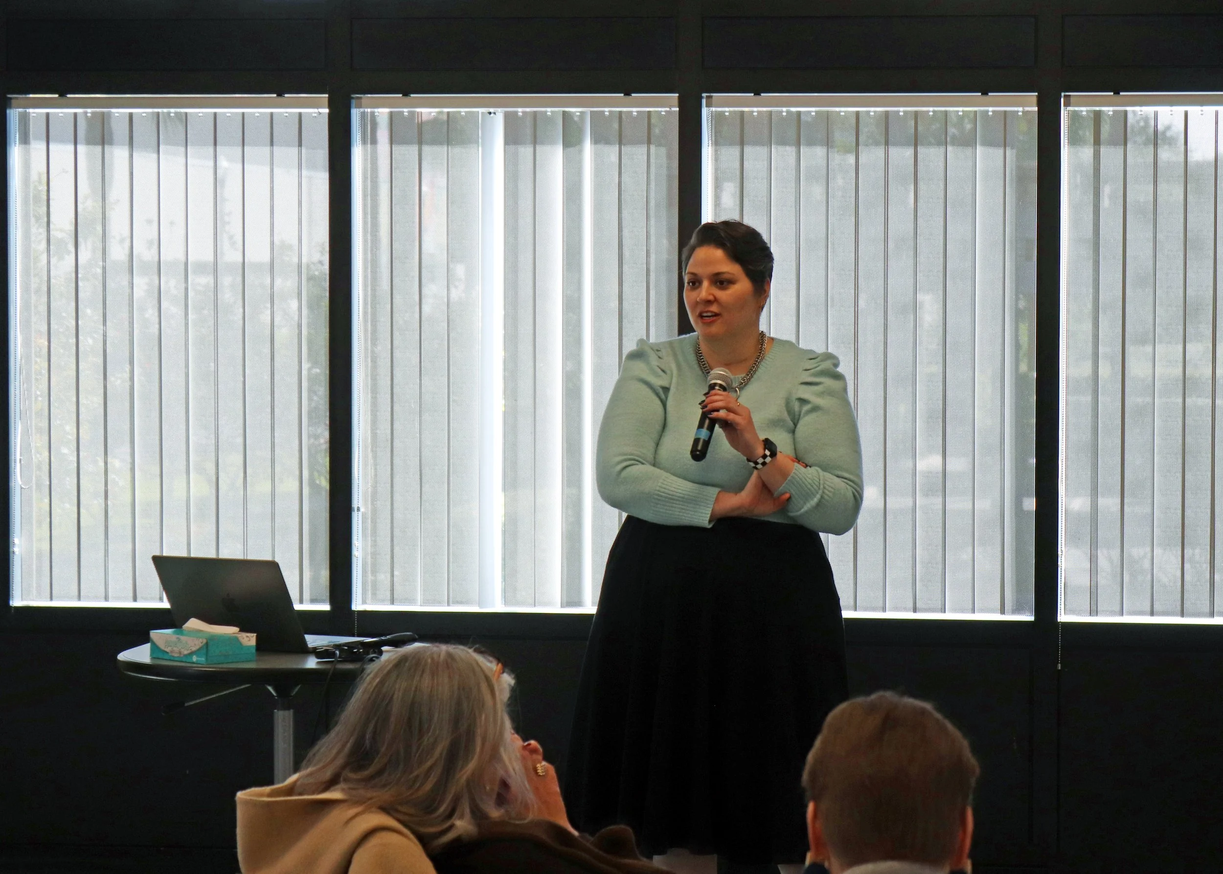 A woman with short dark hair, wearing a light green sweater with puffed shoulders and a chunky chain necklace, is speaking into a microphone in front of large window blinds during a presentation or meeting. Several people are seated in front of her, 