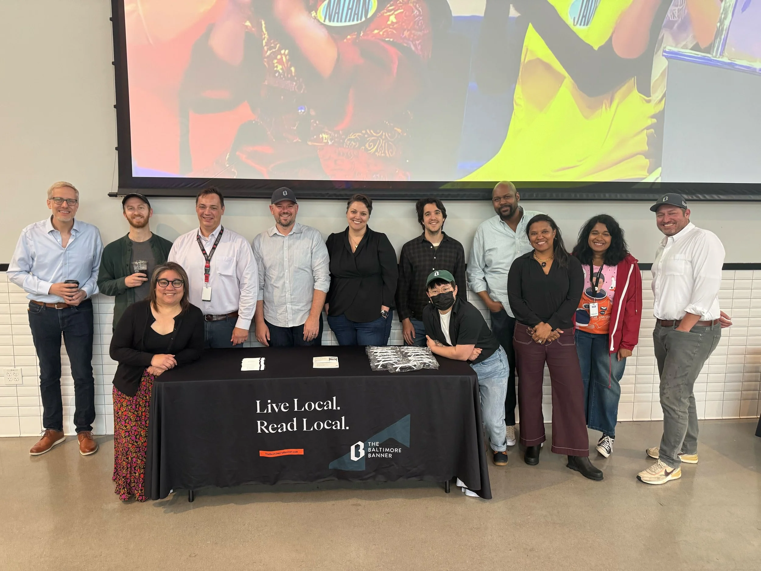 Group of diverse people standing behind a table with a black cloth that reads "Live Local. Read Local" during an indoor event, with a large screen displaying an image behind them.