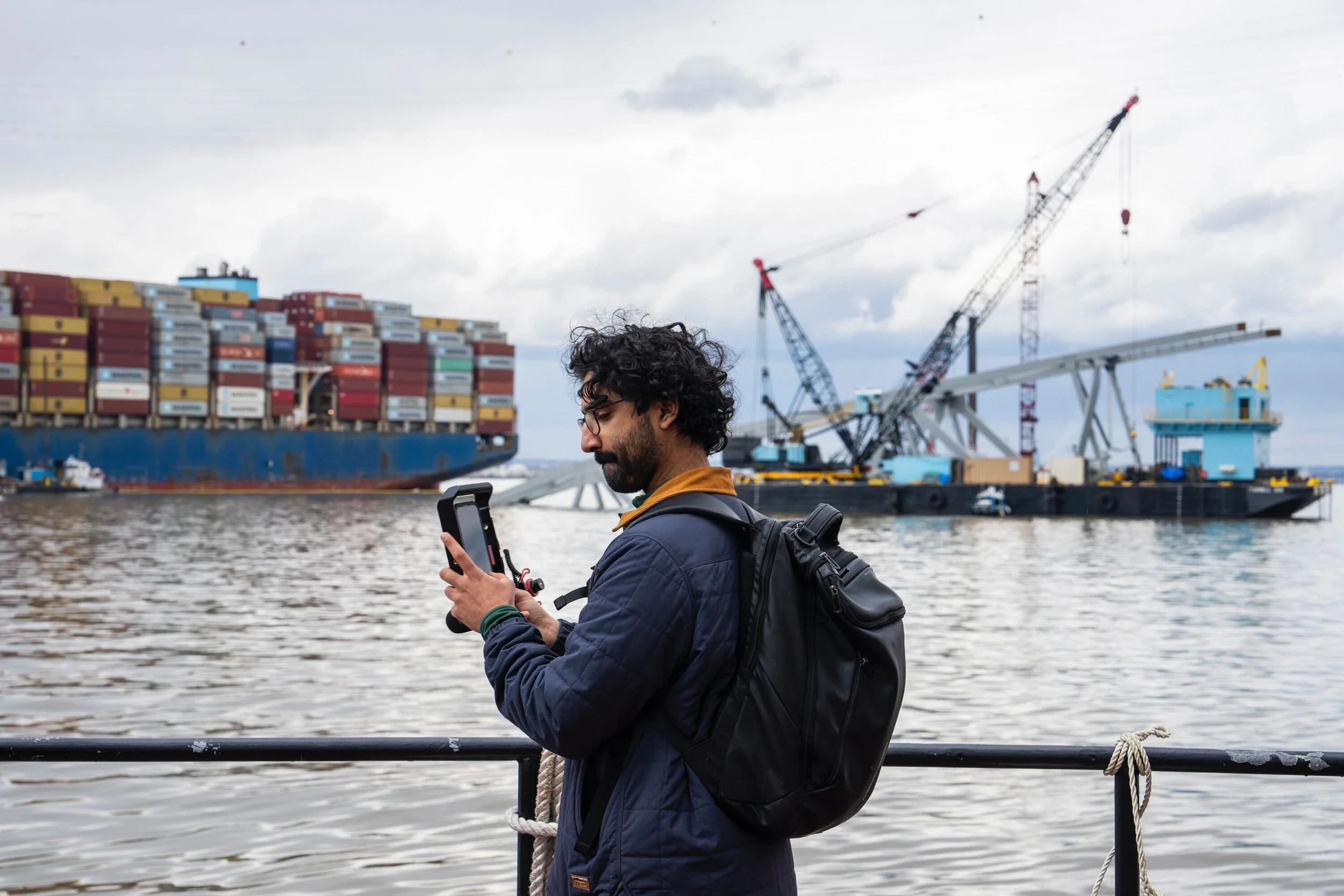 A man with curly hair, glasses, and a backpack stands by water, looking at his phone, with a container ship and cranes in the background.
