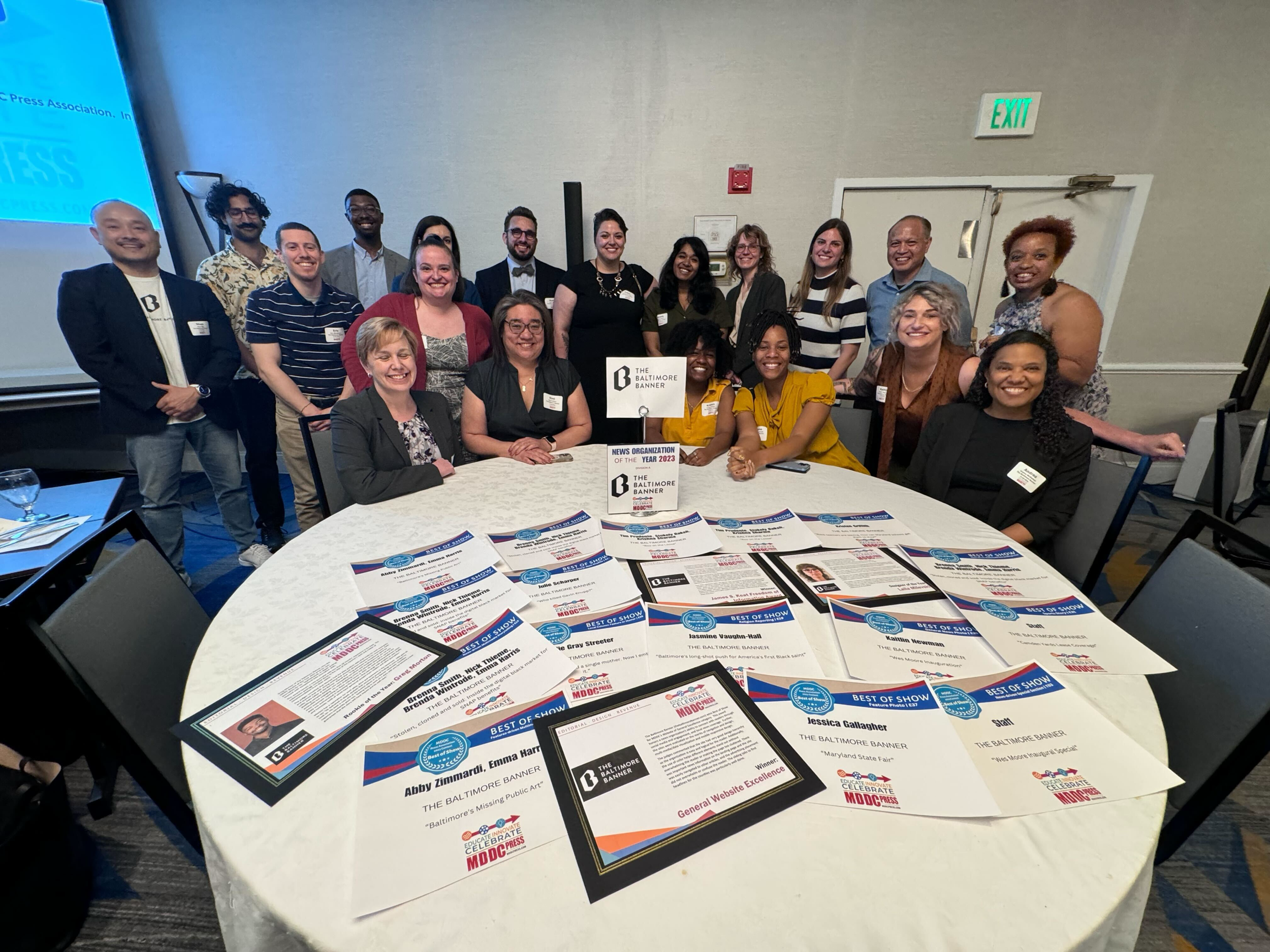 Group of people at a conference, posing for a group photo behind a table with brochures and awards, in a room with beige walls and exit signs.