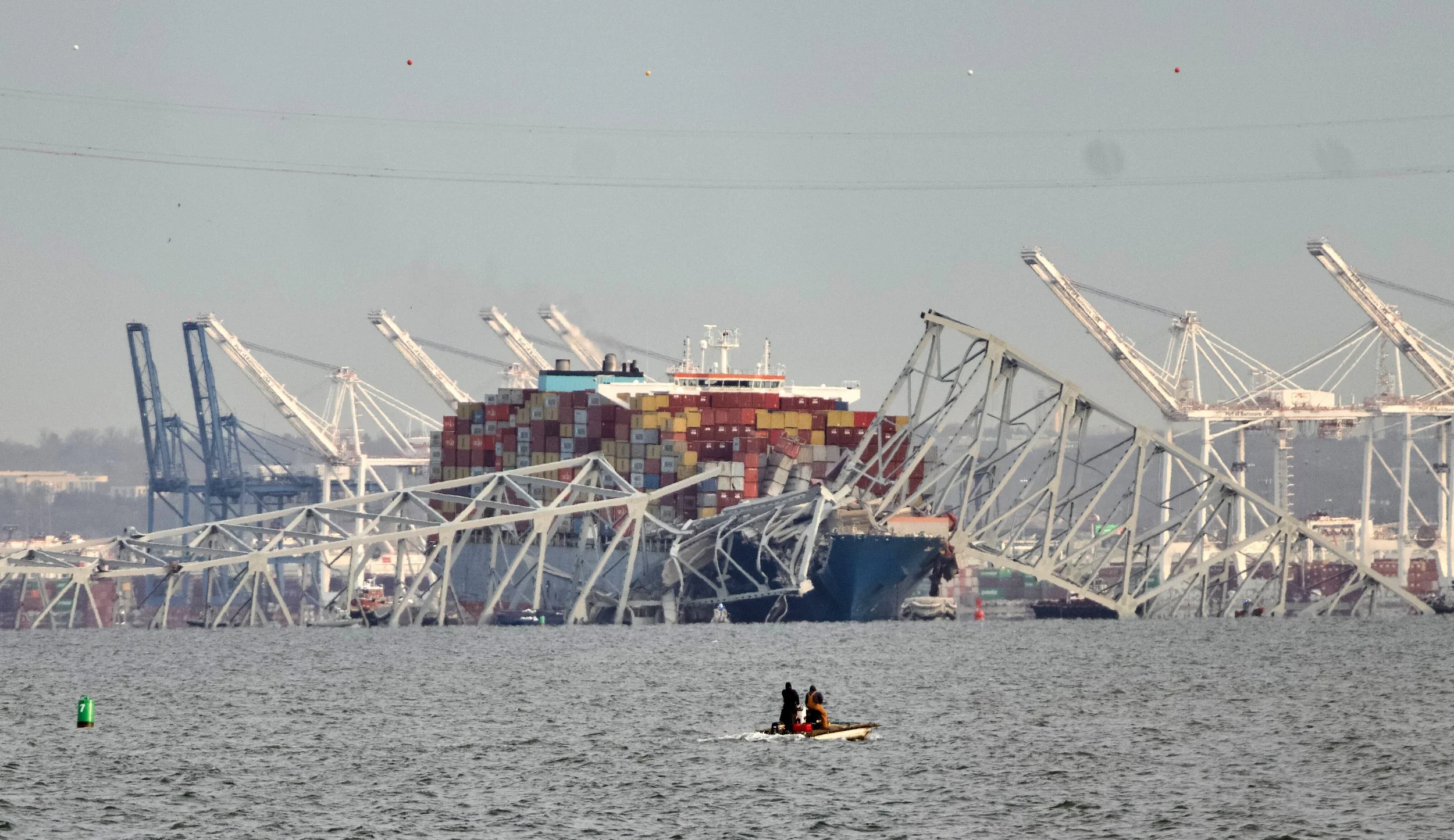A maritime scene with a large cargo ship carrying stacked containers, damaged port cranes, and a boat with two people in the water.